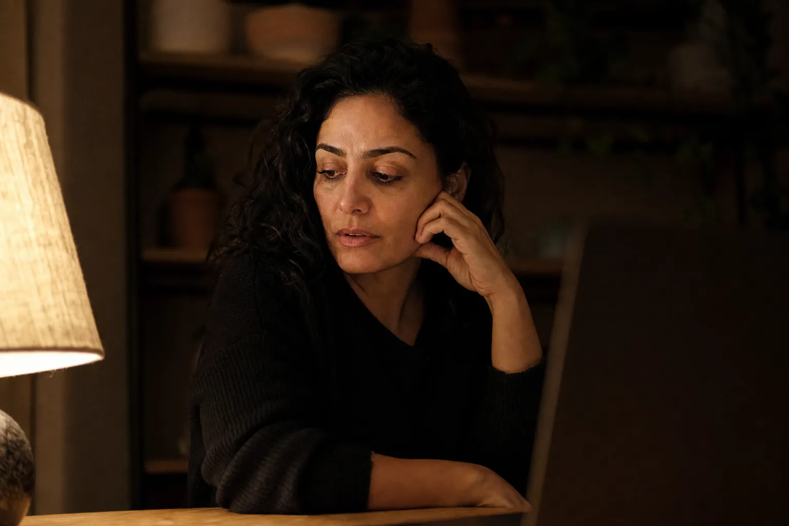A woman at a table in a dim room rests her cheek on her hand and looks down and away from the laptop at the edge of the frame, thinking.